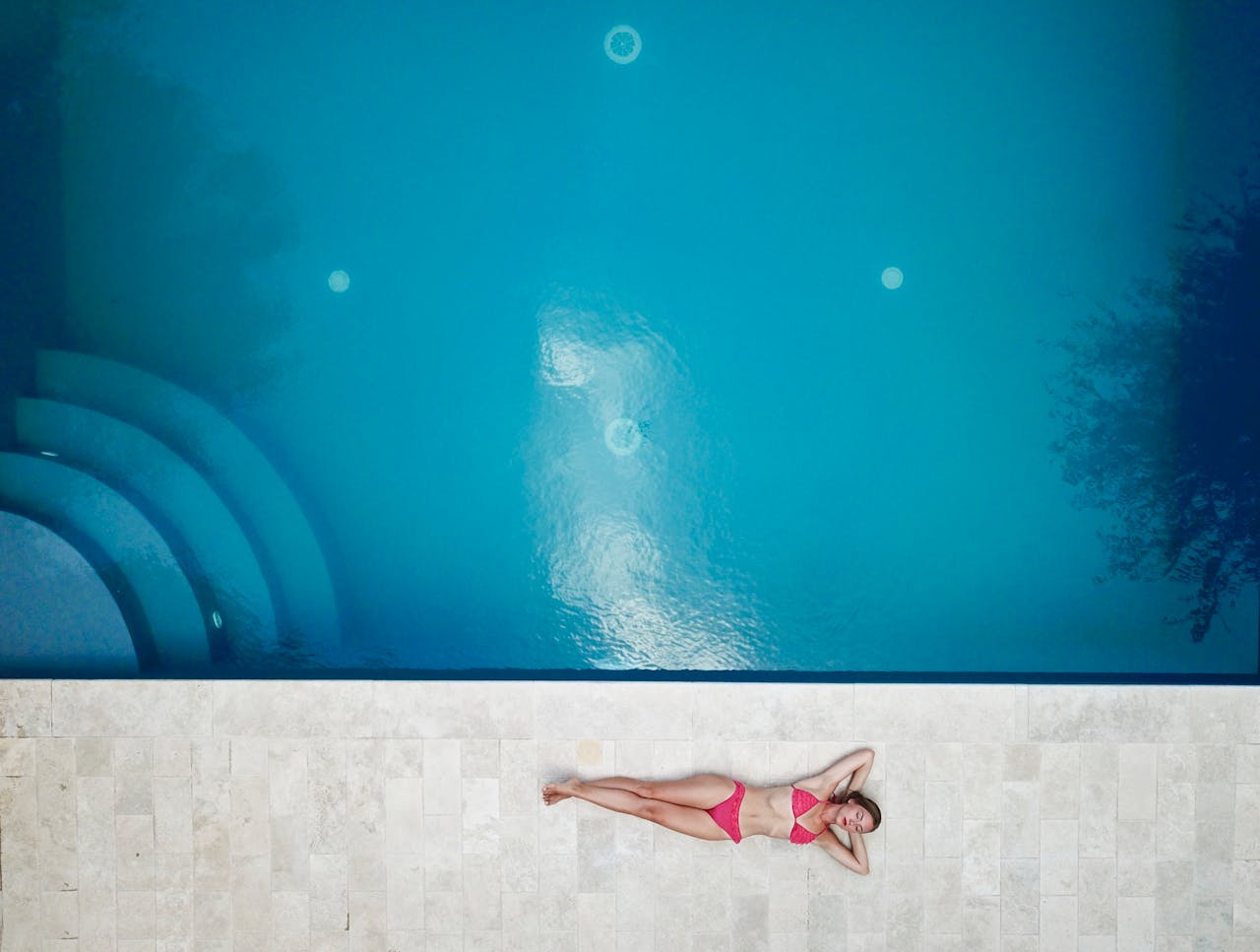 about-bg Bird's-eye view of a woman in a pink bikini relaxing by a bright blue swimming pool.